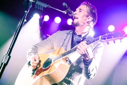 A man playing an acoustic guitar on a stage. He is wearing a grey suit and has a beard. The stage is lit with colorful lights and there is a microphone stand in front of him. The man is singing into the microphone and appears to be in the middle of a performance. He has a focused expression on his face as he plays.