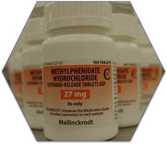 A white medicine bottle with a white cap. The label on the bottle reads "Methylphenidate Hydrochloride Extended-Release Tablets USP 27 mg Rx only". The label also mentions that the medication is available separately to each patient. In the background there are other white medicine bottles with orange labels. The image appears to be a hexagon-shaped label.