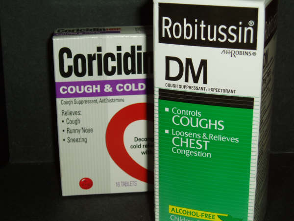 Two boxes of Coricidin and Robitussin DM cough suppressant. The box on the left is white with a red circle in the center and the words "Cough & Cold" written in bold black letters. On the right side of the box there is a green label with the text "Controls Coughs Loosen & Relieves Chest Congestion" in white letters. The label also mentions that the product is alcohol-free and is suitable for children aged 16 and up. The background is black.