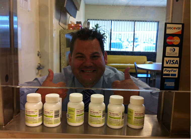 A man standing behind a counter with six white bottles of pills on it. He is smiling and giving a thumbs up with both hands. The man is wearing a blue shirt and has short dark hair. Behind him there is a window with a view of a street and a building. On the right side of the image there are two Visa and PayPal cards. The image appears to be taken in a store or a pharmacy.