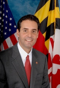 A portrait of a man standing in front of two flags - the United States and the Maryland state flag. The man is wearing a gray suit and a red tie and has a small badge on his lapel. He is smiling and looking directly at the camera. The background is a blue sky with white clouds.