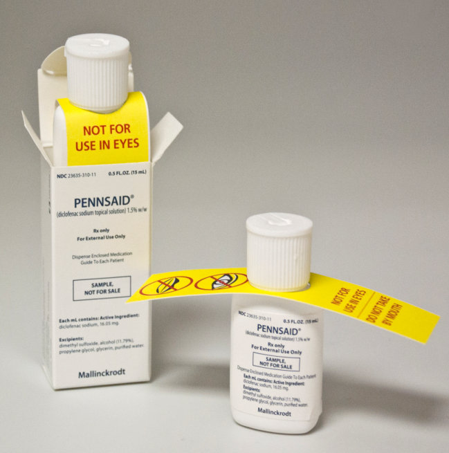 A white box with a yellow label that reads "PENNSAID" and "NOT FOR USE IN EYES". Next to the box there is a small white bottle with a white cap. The label also has a yellow caution tape with a red exclamation mark on it. The box appears to be a prescription bottle for Pennsaid as indicated by the text on the label. The background is plain white.