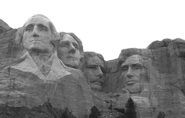 A black and white photograph of the Mount Rushmore National Memorial in South Dakota. It is a large rock formation with four faces carved into it. The faces are of George Washington Thomas Jefferson Theodore Roosevelt and Abraham Lincoln. They are all facing the same direction and appear to be looking off into the distance. The mountain is covered in snow and there are trees and shrubs visible in the background. The sky is overcast and the overall mood of the image is somber.