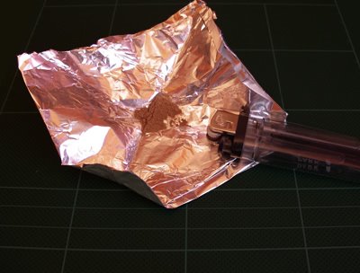 A piece of aluminum foil on a black cutting mat. The foil appears to be crumpled and has a metallic sheen. On the right side of the image there is a small metal object possibly a screw or a screwdriver with a pointed end. The object is resting on the cutting mat and the background is dark.