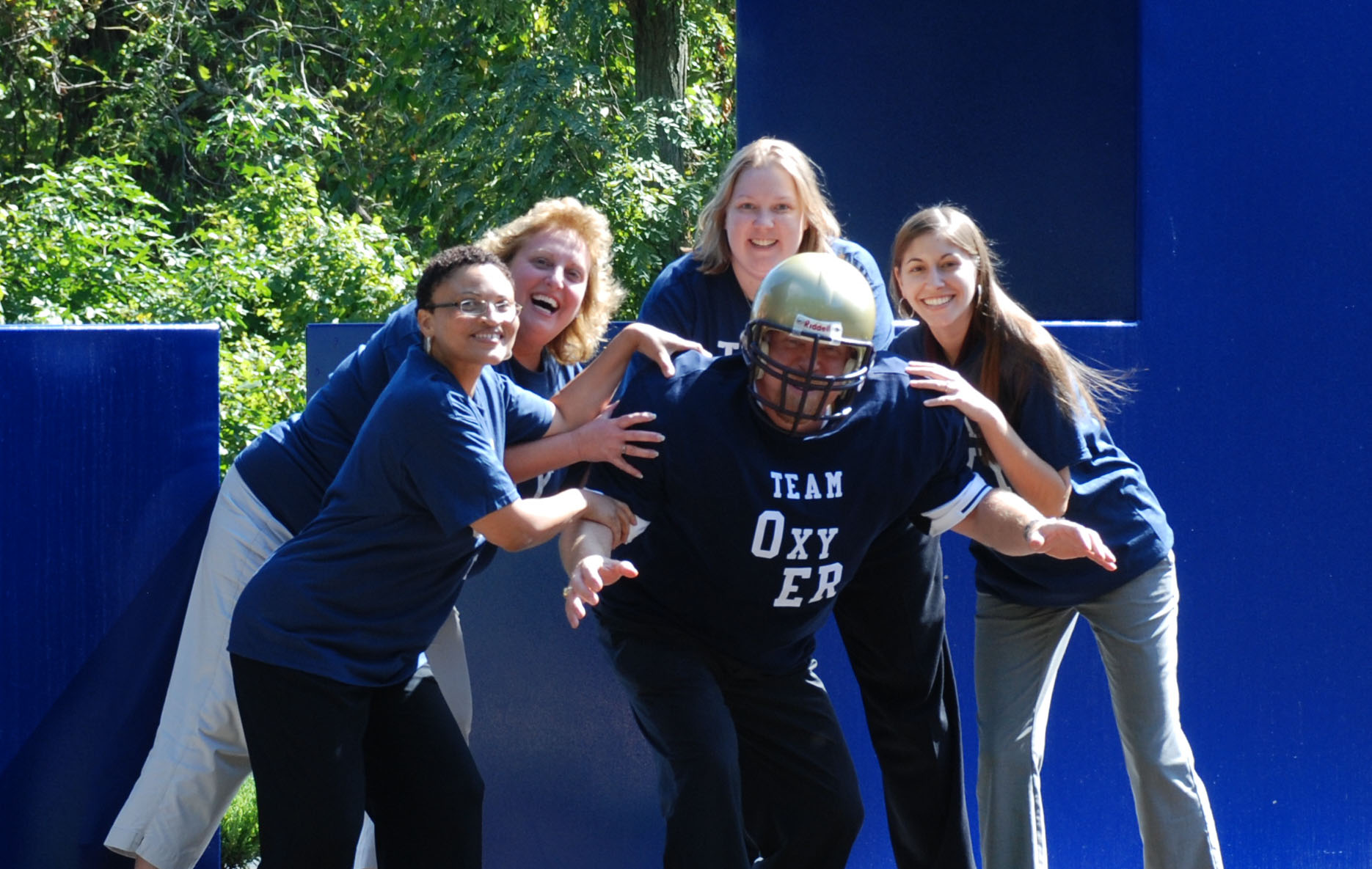 A group of five young women standing in front of a blue wall. They are all wearing blue t-shirts with the words "Team Oxygen" printed on them. The woman in the center is wearing a football helmet and is holding the other woman's arms around her waist. All of them are smiling and appear to be having a good time. The background shows trees and bushes suggesting that the photo was taken outdoors.