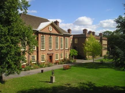A large brick building with a sloping roof and multiple windows. The building is surrounded by a well-manicured lawn and trees. There are two benches on the lawn in front of the building and a small fountain in the center of the lawn. The sky is blue and there are a few clouds in the distance. The overall atmosphere of the image is peaceful and serene.