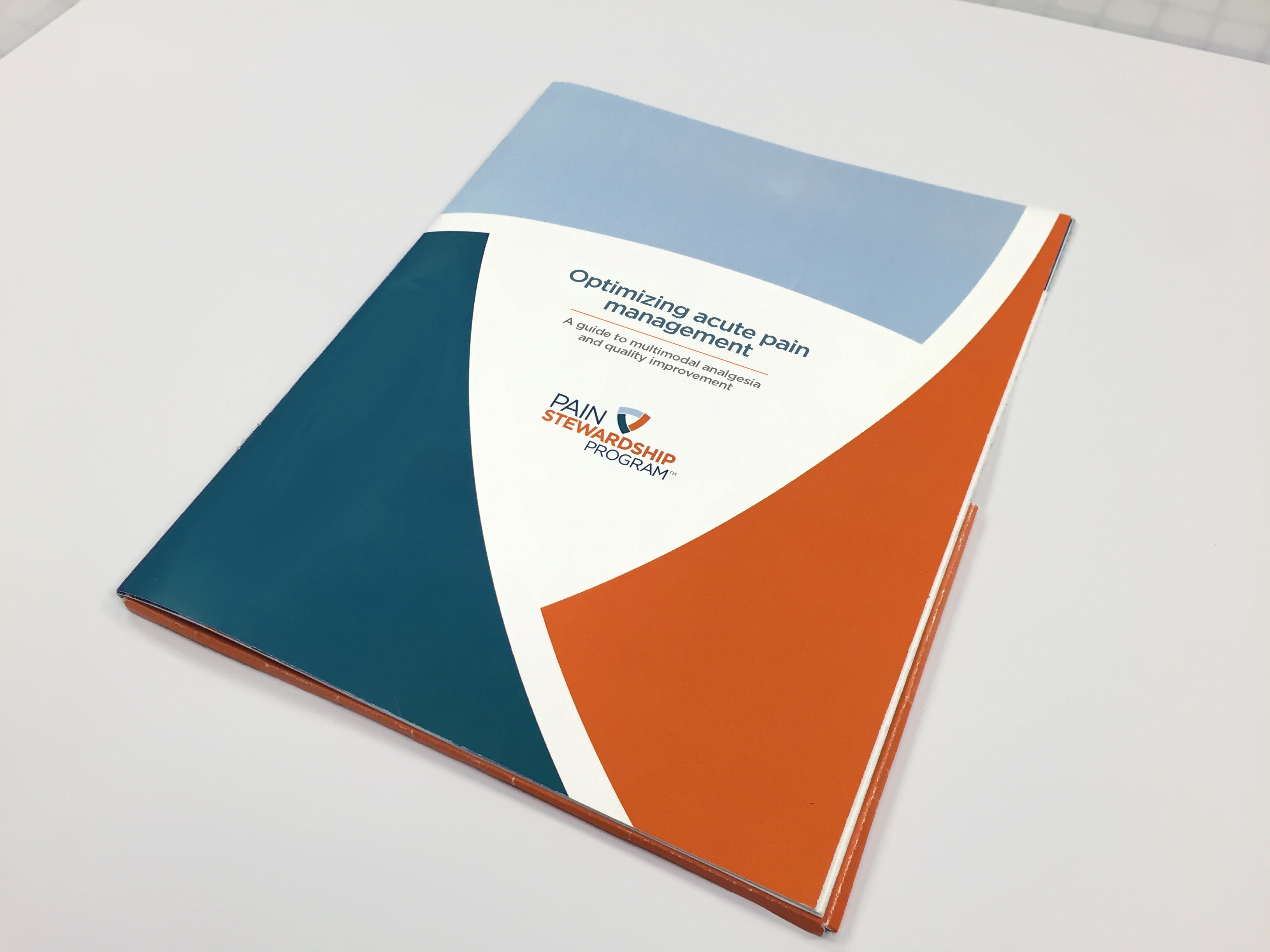 A photograph of a book cover. The cover is rectangular in shape and has a blue and orange color scheme. The top half of the cover has a white background with the title "Optimizing Acute Pain Management" written in bold black font. Below the title there is a logo of a blue circle with a white "P" in the center. The bottom half has an orange and blue wave-like design. The book appears to be lying on a white surface.