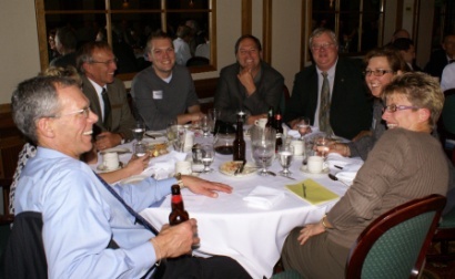 A group of people sitting around a round table in a restaurant. They are all dressed in formal attire and appear to be engaged in a conversation. The table is covered with a white tablecloth and there are plates of food glasses and bottles of beer on the table. The people are smiling and seem to be enjoying each other's company. The restaurant has large windows in the background allowing natural light to enter the space.