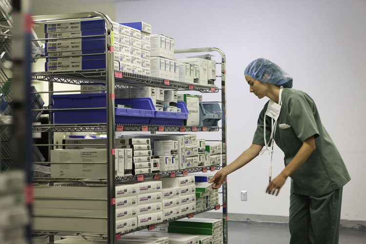 A young woman wearing a green scrub suit and a blue surgical cap on her head. She is standing in front of a large metal rack with multiple rows of boxes and containers on it. The boxes are neatly arranged in rows and the containers are blue and white. The woman is reaching for one of the boxes on the rack and appears to be sorting through them. She has a stethoscope around her neck and is looking down at the boxes. The background is a white wall and the floor is made of concrete.