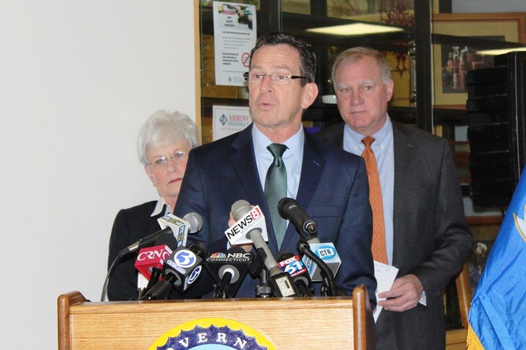 A man standing behind a podium with multiple microphones in front of him. He is wearing a suit and tie and appears to be speaking into one of the microphones. Behind him there are two other people a man and a woman who are also standing behind the podium. The man is holding a piece of paper in his hand and is looking at the camera. On the right side of the image there is a blue flag with the word "Severin" written on it. The background shows a room with a window and a poster on the wall. It appears that the man is giving a press conference.