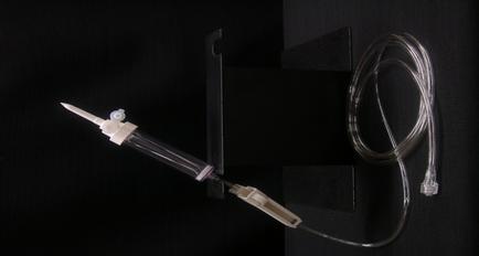 A medical device specifically a syringe with a long thin tube attached to it. The syringe is made of clear plastic and has a white cap on one end. The tube is connected to a black metal bracket on the other end which is likely used to hold the syringe in place. The background is black making the device stand out.