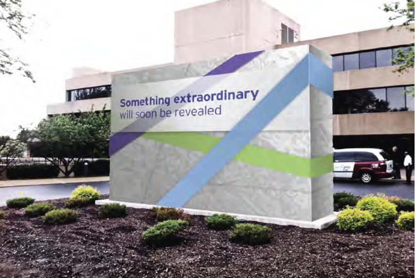 A large concrete sign with a blue and green striped design on it. The sign reads "Something extraordinary will soon be revealed" in bold white letters. The background of the sign is a modern building with large windows and a parking lot with a white car parked in front. The sky is overcast and there are trees and bushes in the foreground.