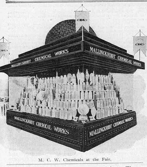 A black and white illustration of a display stand for M.C.W. Chemicals at the Fair. The stand is made of wood and has a dome-shaped roof with a chimney on top. The front of the stand has a sign that reads "Mallinckrodt Chemical Works" in bold letters. Below the sign there is a large display of glass bottles arranged in a pyramid-like structure. The bottles are of different sizes and shapes with some being tall and others being short. There are also several smaller bottles on the stand and a banner on the right side of the image. The background is plain white.