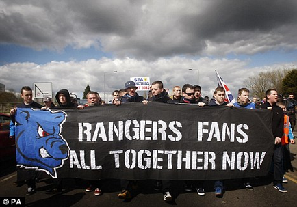 A group of people holding a large banner that reads "Rangers Fans All Together Now". The banner is black with white text and a blue logo of a blue bear on the left side. The people are standing on a street with a cloudy sky in the background. They appear to be participating in a protest or rally. Some of the people are holding flags and banners while others are holding signs and banners. The overall mood of the image is one of solidarity and support for Rangers fans.