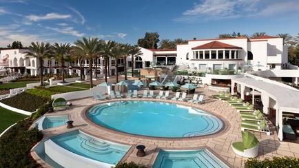 An aerial view of a luxurious resort with a large swimming pool in the center. The pool is surrounded by a circular patio with lounge chairs and umbrellas providing a perfect spot to relax and enjoy the view. On the right side of the image there is a large white building with a red roof which appears to be a hotel or villa. The building has multiple floors and balconies and there are palm trees and other tropical plants surrounding it. The sky is blue with a few clouds and the overall atmosphere is peaceful and inviting.