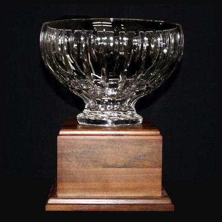 Of a glass bowl on a wooden base. The bowl is round and has a wide base with a narrow neck and a wide opening at the top. It appears to be made of clear glass with a ribbed pattern on the sides. The base is square in shape and is made of a dark wood with a smooth finish. The background is black making the bowl stand out.
