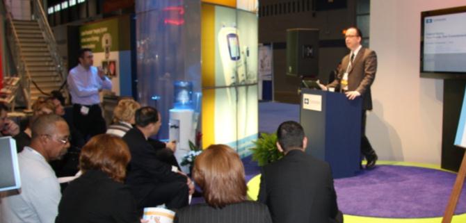 A man standing at a podium in front of a crowd of people. He is speaking into a microphone and appears to be giving a presentation. The crowd is seated in a semi-circle around the podium attentively listening to the speaker. On the right side of the image there is a large screen displaying a presentation slide. In the background there are stairs leading up to a higher level of the room. The room is decorated with colorful lights and plants.