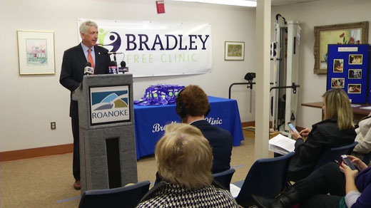 A man standing at a podium and speaking into a microphone. He is wearing a suit and tie and appears to be giving a speech. Behind him there is a banner that reads "Bradley Free Clinic" and a table with a blue tablecloth. There are several people sitting in chairs in front of the podium attentively listening to the man speak. On the right side of the image there are several framed pictures hanging on the wall.