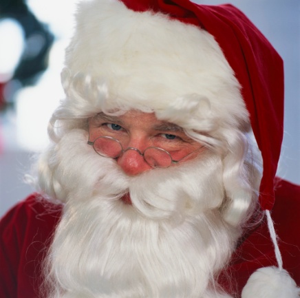 A close-up portrait of Santa Claus. He is wearing a traditional red suit with a white fur trim and a red hat with a gold trim. He has a long white beard and mustache and is wearing round glasses. His eyes are blue and he has a serious expression on his face. The background is blurred but it appears to be a room with a Christmas tree and other decorations.