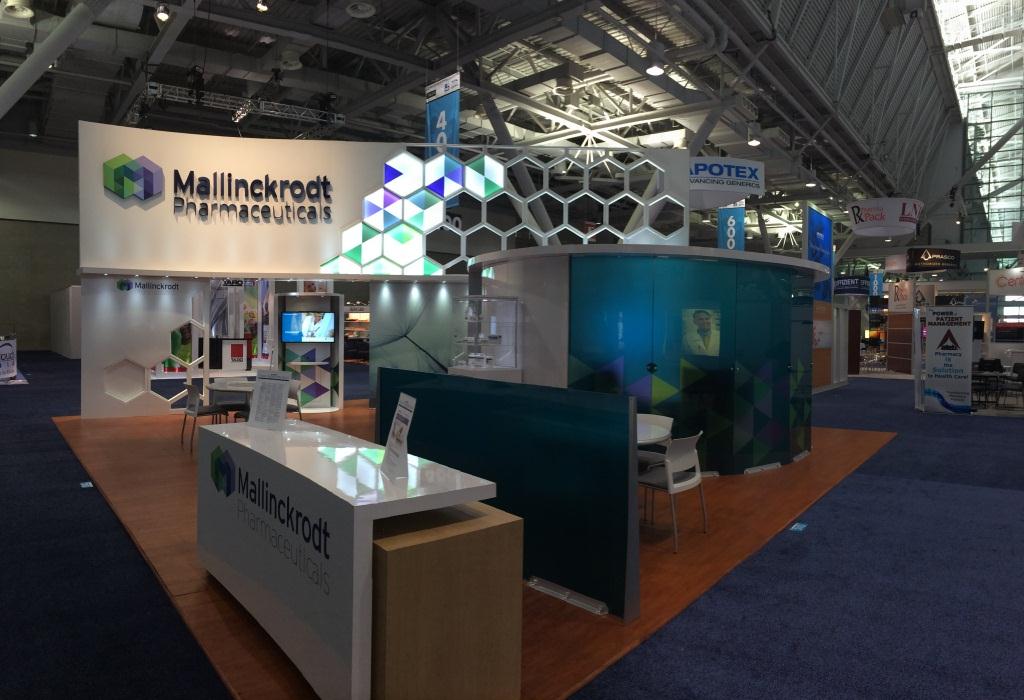 A trade show booth with a modern design. The booth is located in a large hall with a high ceiling and large windows. The walls of the booth are decorated with a colorful geometric pattern in shades of blue green and white. There is a large banner on the left side of the image that reads "Mallinckrodt Pharmaceuticals" in bold letters. On the right side there is a curved booth with the company's logo on the front and a desk in front of it. In the background there are several other booths and banners with information about the company. The floor is covered with a blue carpet.