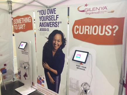Three banners hanging on a white tent. The first banner on the left has a photo of a woman with long dark hair and a big smile on her face. The second banner has text that reads "You owe yourself answers!" and "Jessica D." The third banner has the text "Curious?" and the logo of glenda (Fingolimod) on the top right corner. The banners also have a small screen attached to one of the banners. The background of the image appears to be an exhibition or trade show booth.