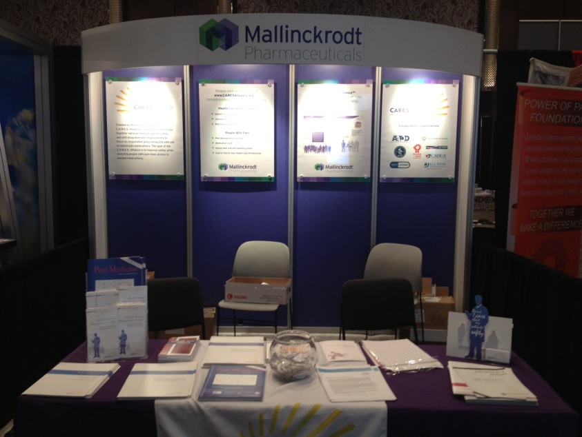 A booth at a trade show or exhibition. The booth is called "Mallinckrodt Pharmaceuticals" and has a purple and white color scheme. It has a large banner at the top with the company's logo and contact information. Below the banner there are three panels with information about the company. <br /><br />The panels are arranged in a triangular formation with the largest panel on the left and the two smaller panels on the right. Each panel has a different image and text on it.<br /><br />In front of the booth there is a table with a purple tablecloth and a white tablecloth. On the table there has a few items such as brochures pamphlets and a small figurine of a person. There are also a few chairs in the background. The overall atmosphere of the image is professional and organized.