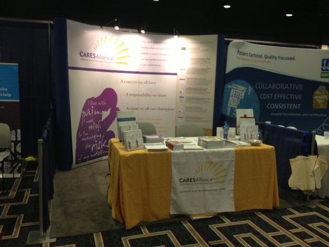 A trade show booth with a table in the center. The table is covered with a yellow tablecloth and has a banner that reads "CARESAlliance" in white letters. On the table there are various items such as books pamphlets and a water bottle. Behind the table is a large banner with a purple background and white text. The banner also has a yellow sunburst design and the words "Collaborative Cost Effective Consistent" in blue letters. There are also two smaller banners on either side of the table. The booth is set up in a large room with a carpeted floor and a blue curtain in the background.