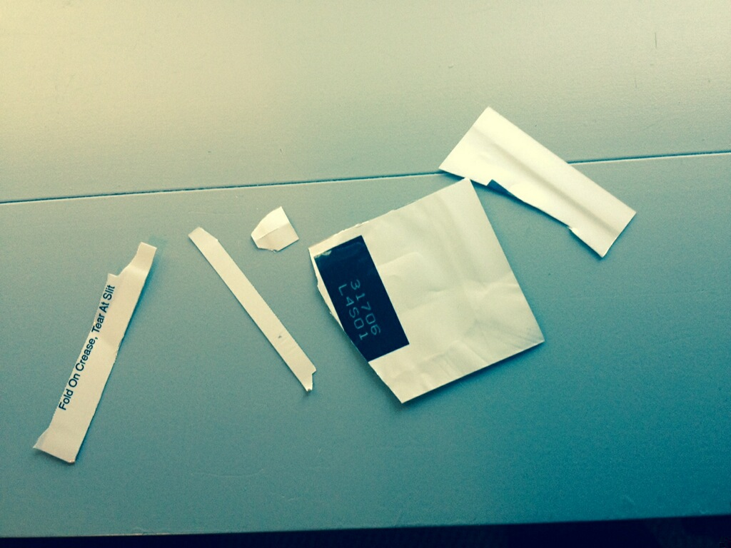 A white envelope with a black label on it lying on a light blue surface. The envelope appears to be made of paper and has a rectangular shape with a flap on top. There are several pieces of the envelope scattered around it some of which are torn off and some are lying on the surface. On the left side of the image there is a piece of white paper with a label that reads "Made in China". The background is a plain white wall.