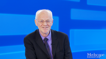 A portrait of an elderly man in a suit and tie. He is standing in front of a blue background with a grid pattern. The man is bald and has a friendly expression on his face. He appears to be in his late 60s or early 70s and is wearing glasses. He has a microphone attached to his lapel and is looking directly at the camera. The image is likely from a news or interview setting.