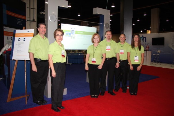 A group of six people standing in front of a large screen in a trade show booth. They are all wearing green shirts and black pants and are smiling at the camera. Behind them there are several other booths and banners with information about the trade show. On the left side of the image there is a sign with a blue background and white text. The floor is covered in a red carpet and there are pillars in the background.