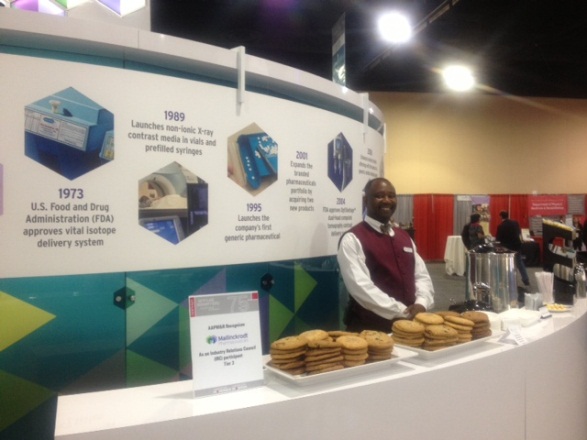 A man standing behind a counter at a trade show. The counter is white and has a blue and green design on it. On the left side of the counter there is a large banner with information about the U.S. Food and Drug Administration (FDA) and its delivery system. The banner also mentions that the FDA approved the FDA's approval of a new delivery system for food and drug delivery systems.<br /><br />In the center of the image the man is standing behind the counter and is smiling at the camera. He is wearing a maroon vest and a white shirt. Behind him there are other booths and people walking around. The background is blurred but it appears to be an indoor space with a high ceiling and bright lights.