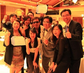 A group of people posing for a photo in a banquet hall. There are nine people in the photo all of whom are smiling and giving a thumbs up. They are all dressed in formal attire with the men wearing suits and the women wearing dresses. Three are holding certificates or awards and one is holding a metal plate possibly some sort of trophy. The group is standing in front of a table with a white tablecloth and there are other tables and chairs in the background. The room appears to be well-lit with chandeliers hanging from the ceiling.