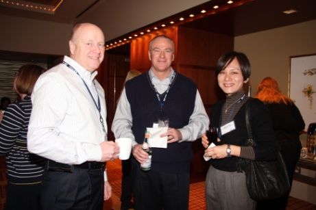 Three people standing in a room with a high ceiling and wooden walls. They are all smiling and appear to be engaged in a conversation. On the left is an older man wearing a white shirt and black pants holding a white coffee cup in his left hand. In the middle is a middle-aged man with short blonde hair and a blue vest and on the right is a young woman wearing a black top and gray pants. She is also holding a drink in her right hand. All three people are wearing name tags around their necks. There are other people in the background but they are not visible.