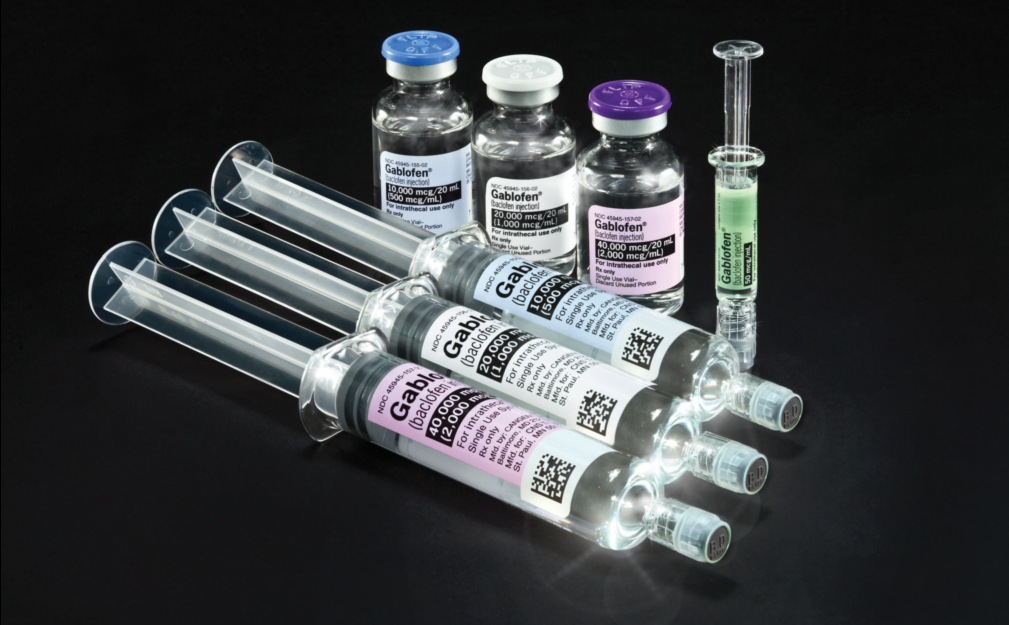 A group of six syringes of different sizes and shapes arranged in a row on a black background. There are six vials of different colors - blue white pink purple and green - with labels on them. The vials have blue caps and are filled with a clear liquid. The syringe on the right side of the image has a small amount of green liquid in it which appears to be a test tube. The background is also black making the colors of the vials stand out.