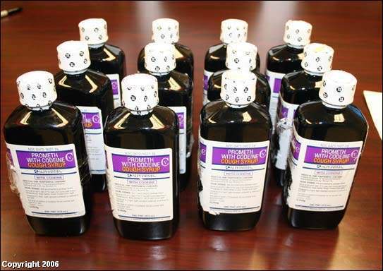 A group of 12 bottles of liquid arranged in a row on a wooden table. The bottles are black in color and have white caps. Each bottle has a purple label with white text that reads "Prometh with Codeine Cough Syrup". The bottles appear to be filled with a dark liquid possibly cough syrup. 