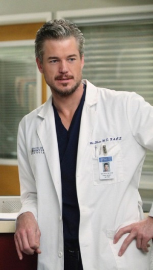 A man in a white lab coat standing in a hospital room. He is wearing a navy blue scrub top and has a name tag attached to his left chest pocket. He has a serious expression on his face and is looking directly at the camera. The background shows a window with blinds and a desk with a computer monitor. The man appears to be a doctor or a medical professional.