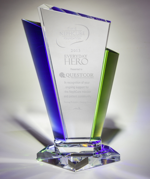 Of a glass award. The base is diamond-shaped with a green and blue gradient on the sides. On the top of the base there is a white rectangular plaque with the words "2013 Everyday Hero" and "Questcor" written in black text. Below the plaque it reads "In recognition of your outstanding support for the Nephecure Foundation and patient community." The plaque is placed on a white reflective surface. The background is blurred making the award the focal point of the image.