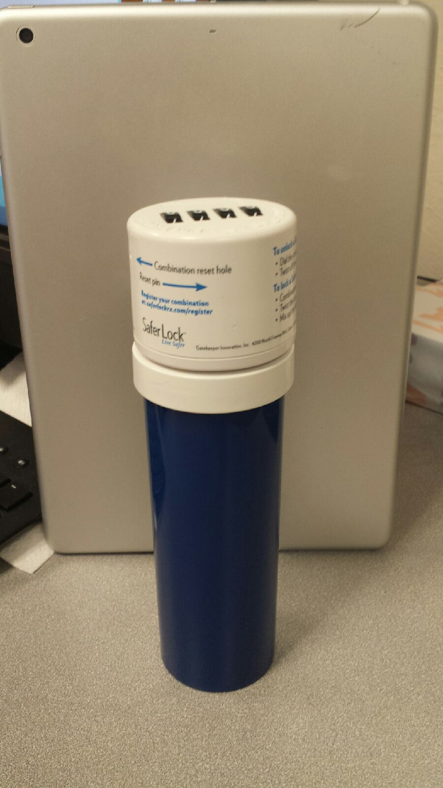 A photograph of what appears to be a blue pill bottle with a cap that is activated by a combination lock. The cap has the logo for SaferLock on it. In the background there is a laptop and a keyboard suggesting that the image was taken in an office or workspace.