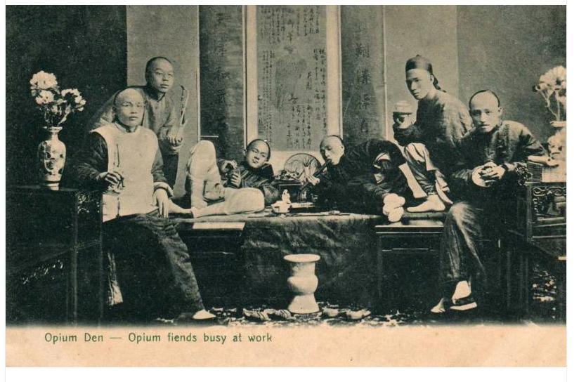 A black and white photograph of a group of six men sitting around a table in a room. The men are dressed in traditional Chinese clothing. The table is covered with a tablecloth and there are vases of flowers on either side of the table.<br /><br />In the center of the image there is a man sitting on a chair with his legs crossed and a serious expression on his face. He is wearing a long robe and a hat with a feather on top. To his left there are two men standing behind him one of them is holding a book and the other is looking at the camera. To the right another man is sitting at the table with a cup of tea and a plate of food in front of him. In the background there appears to be a window with curtains and a chandelier hanging from the ceiling. The photograph appears to have been taken in the early 20th century. The caption on the image reads "Opium Den - Opium fiends busy at work."