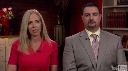 A man and a woman sitting in a living room. The woman is on the left side of the image wearing a red dress and has blonde hair. She is looking directly at the camera with a serious expression on her face. The man on the right side is wearing a gray suit and a pink tie. He has a mustache and appears to be in his late 40s or early 50s. Behind them there is a bookshelf with books and a vase of flowers. The background is a brick wall.