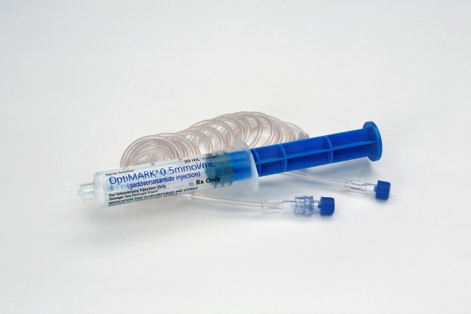 A medical syringe with a blue handle and a clear plastic tube. The syringe is lying on a white background. The tube is coiled up and has a label on it that reads "Optimark 0.5mm/ml". 