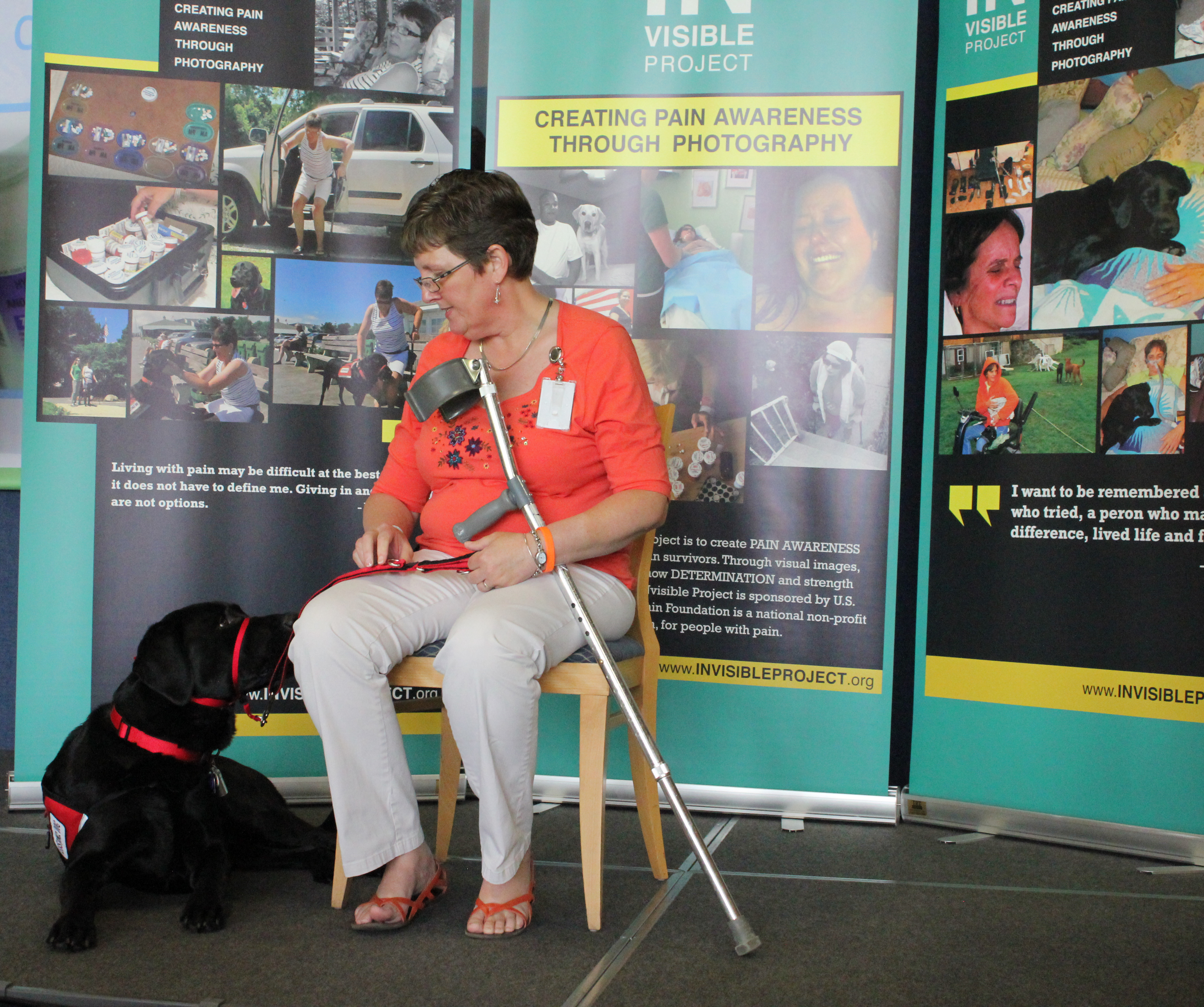 A woman sitting on a chair with a black dog on a leash. The dog appears to be a service animal due to its red vest and leash. The woman is wearing an orange shirt white pants and orange sandals. She is holding a crutch. Behind her there trifold presentation with images and text related to creating pain awareness through photography. The banners are blue and yellow in color and have images of people and animals on them.