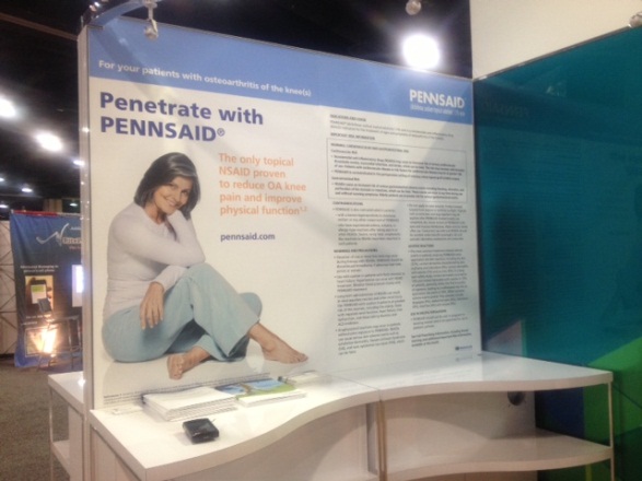 Of a booth at a trade show. The booth has a blue and white color scheme with a large banner on the left side that reads "Penetrate with Pennsaid". On the right side of the banner there is an image of a woman sitting on the floor with her legs crossed and a smile on her face. She is wearing a white shirt and blue pants. Below the image there are several brochures and information about the product. The background of the booth is a white wall with a green and blue design.