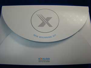 A white envelope with a circular logo on the front. The logo is a grey circle with an X in the center and the words "New Beginning Kit" written around it. The envelope appears to be new and unused with no visible damage or wear. The background is dark making the white envelope stand out. On the bottom right corner of the envelope there is a blue logo that reads "EXALGO".