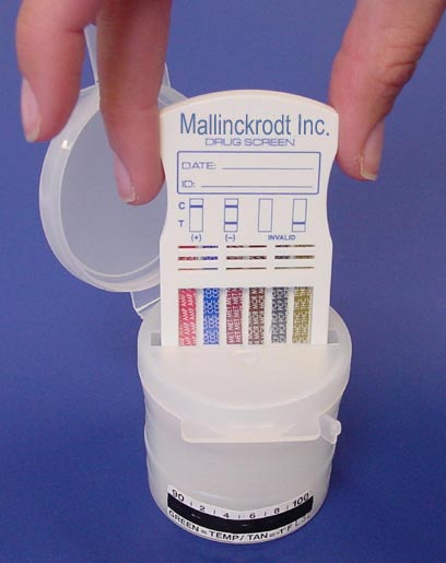 A person's hand holding a small white pill bottle with a lid. The bottle is labeled "Mallinckrodt Inc. Drug Screen" and has a label on it that reads "Date". The label also has a barcode and some text on it. The pill bottle appears to be empty and the lid is open revealing the contents inside. The background is a plain blue color.