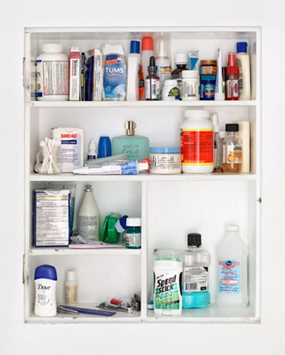 A white medicine cabinet with three shelves. The cabinet is open and the contents are neatly arranged on the shelves. On the top shelf there are several bottles of different sizes and colors. The bottom shelf has a few items on it including a blue bottle of deodorant a white box of toothpaste a green bottle of lotion and a blue container of toothbrushes. The items are arranged in a neat and organized manner and the cabinet appears to be mounted on a white wall.