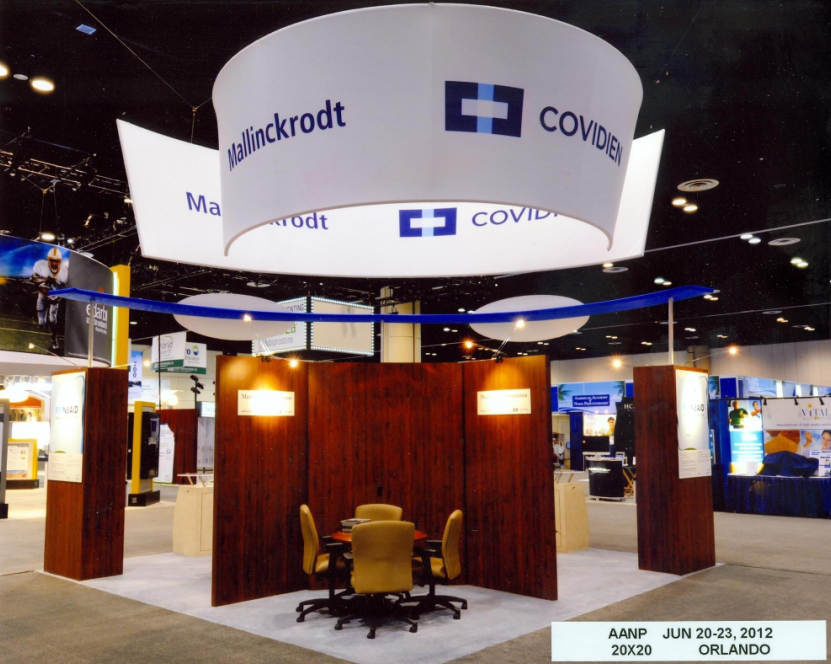 A trade show booth with a large white circular banner hanging from the ceiling. The banner has the logo of Mallinckrodt and Covidien both of which are blue and white. Below the banner there is a wooden booth with two yellow chairs and a table. The booth is located in an exhibition hall with other trade show booths in the background. The floor is covered with a gray carpet and there are banners and signs on the walls. The overall atmosphere of the booth is modern and professional.