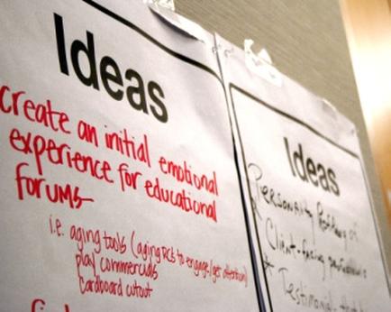 Two whiteboards attached to a bulletin board with clips. On the left board there is a list of ideas written in red marker. The list is titled "Ideas" and "Create an initial emotional experience for educational forums". The list includes: - I.e. aging tools (aging tools) - Play commercials - Cardboard cutout - Personalization - Testimonial The whiteboards appear to be part of a larger presentation or brainstorming session. The background is blurred but it seems like the focus is on the whiteboards.