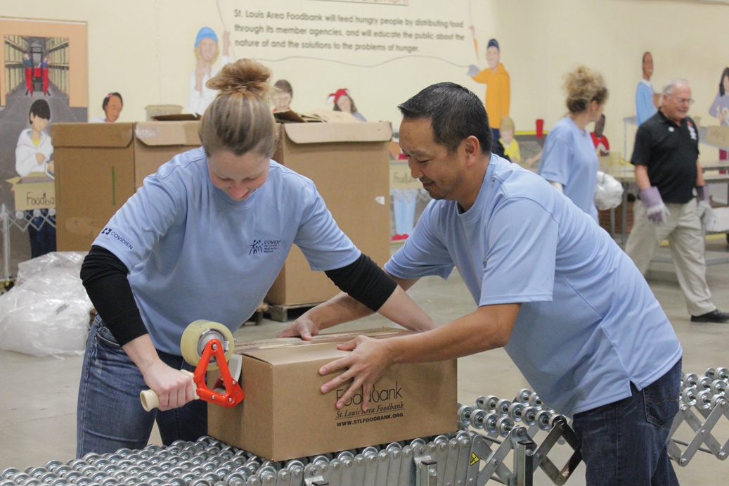 A man and a woman in a warehouse. They are both wearing blue t-shirts and are working together to assemble a cardboard box on a conveyor belt. The woman is holding a pair of scissors and appears to be cutting the box with it while the man is holding it with both hands. In the background there are several other people working in the warehouse as well as cardboard boxes and other items scattered around. On the wall behind them there is a mural of a group of people and a sign that reads "Food Bank."