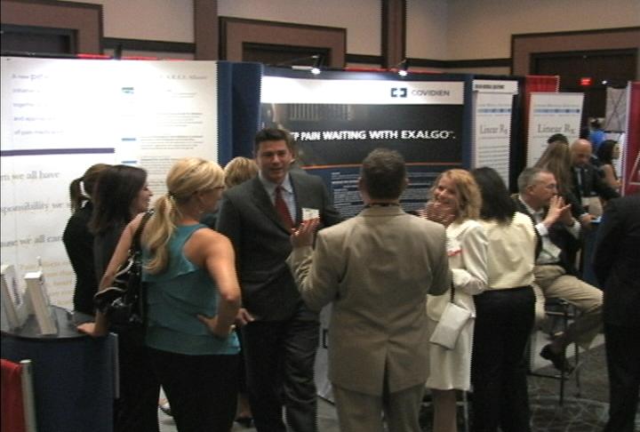 A group of people gathered around a poster board at an exhibition. The poster board has text and images on it and there are several people standing in front of it. The people appear to be engaged in conversation with some of them looking at the poster and others gesturing with their hands. The background of the image is a large room with a high ceiling and a chandelier hanging from the ceiling. It appears to be a conference or event space.